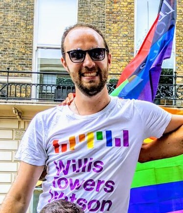 A man wearing a white T-shirt and sunglasses with a pride flag behind him