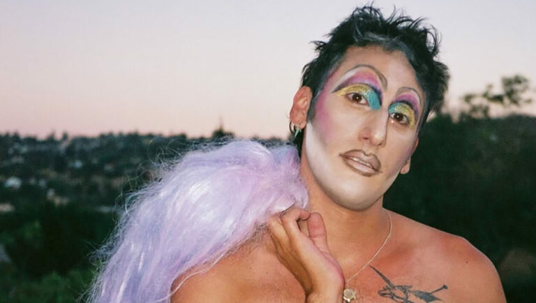 A person holding a purple wig over their shoulder looking into the camera