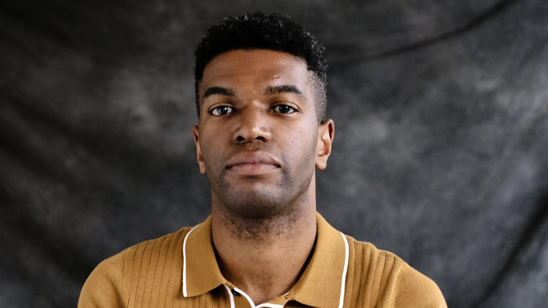 Okechukwu Nzelu sat against a misty grey background wearing a mustard top.