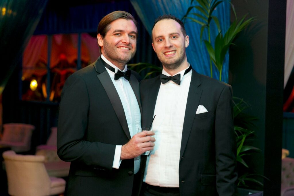 Two men wearing tuxedos smile into the camera