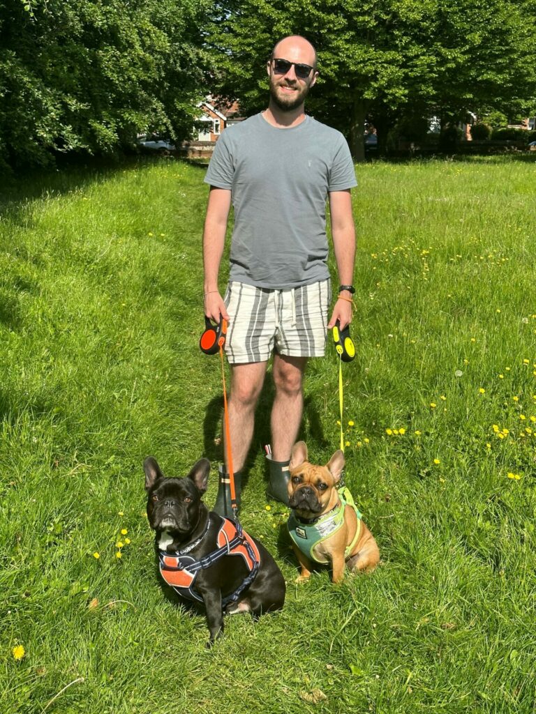 A man wearing a grey top and striped shorts stands in grass holding two dogs by their leashes