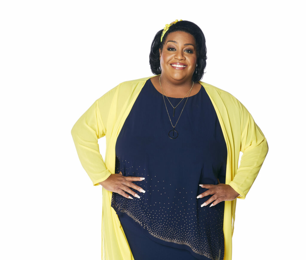 Alison Hammond stands with her hands on her hips, wearing a black dress and yellow shawl