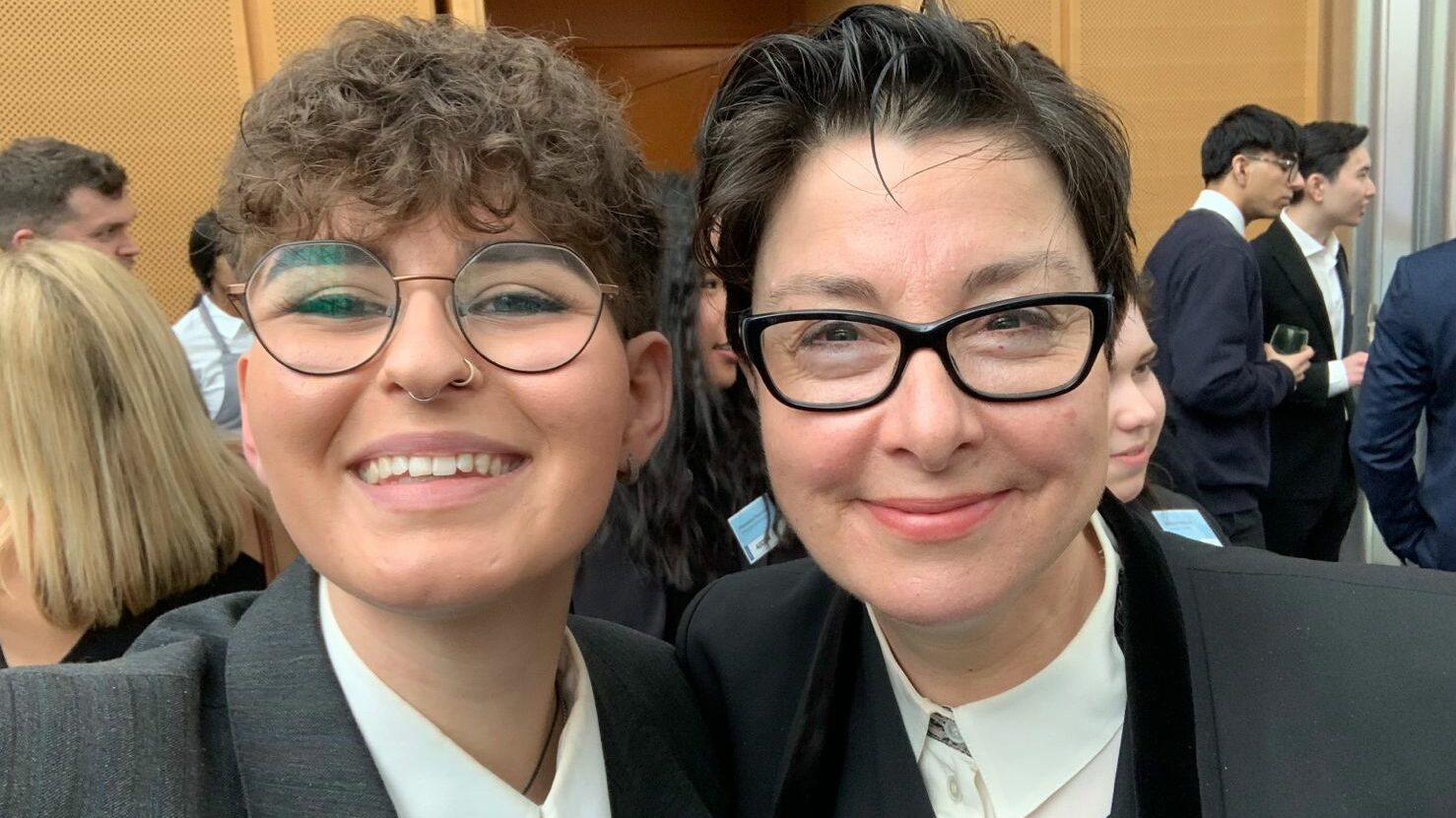 Two people wearing suits and glasses hug and smile into the camera