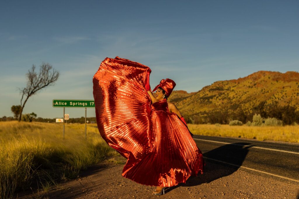 A dra queen in a red dress standing next to a sign saying Alice Springs