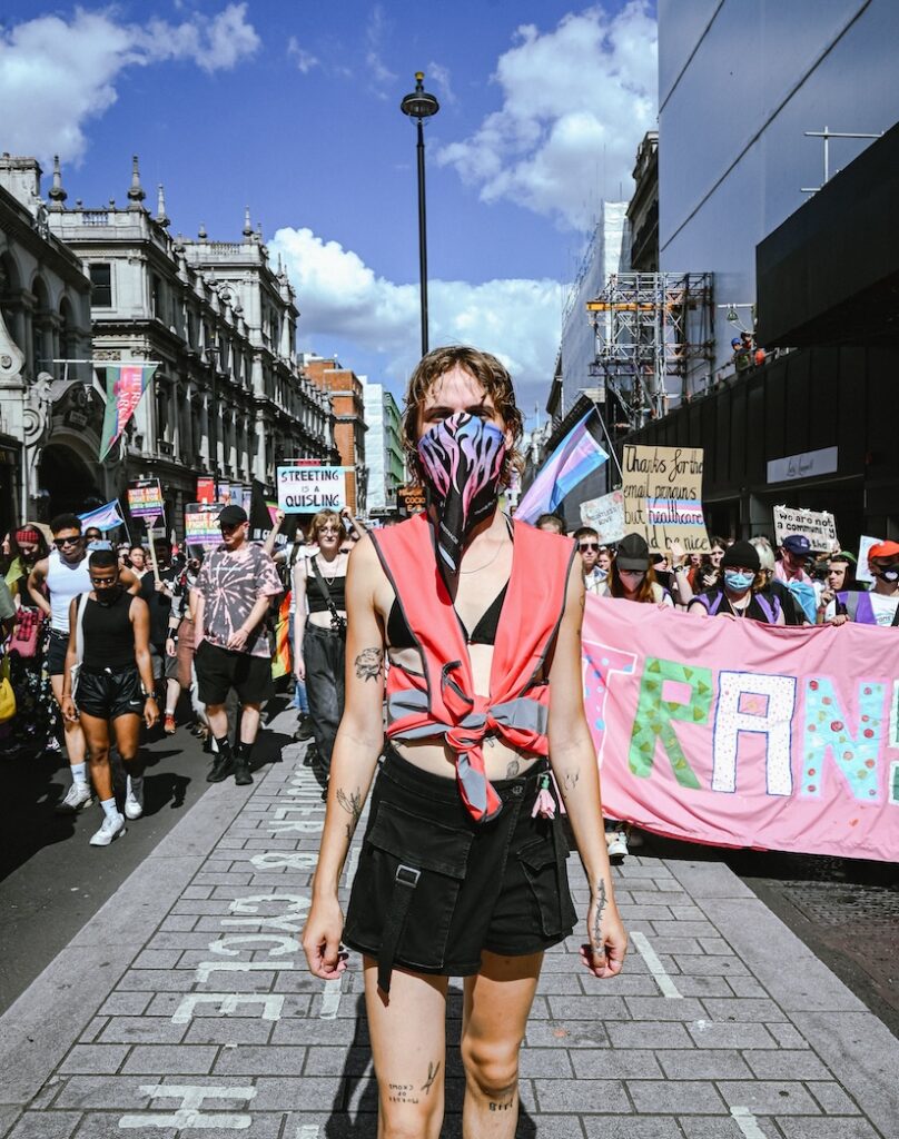 A crowd shot, centred on a person wearing a face mask with fire in trans flag colours