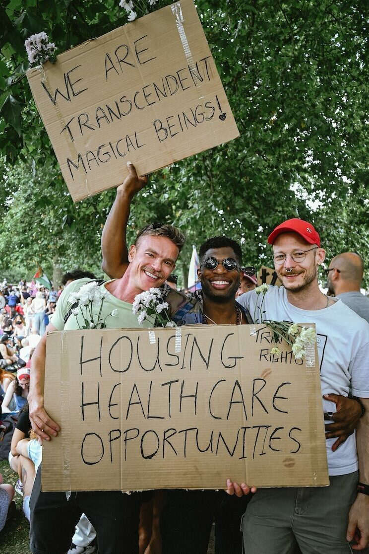 Three friends pose in Hyde park, holding signs saying 'we are transcendent human beings' and 'housing healthcare opportunities'