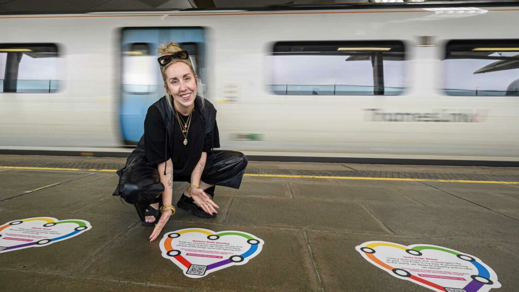 A person kneels down over a Pride heart sign inside a train station