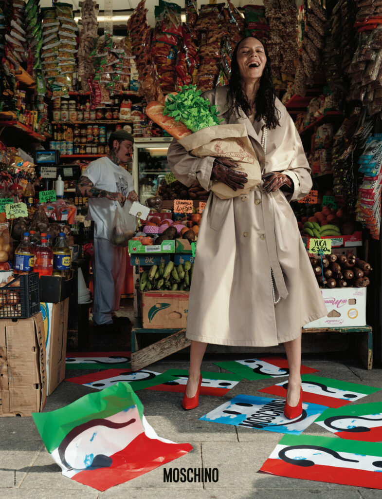 A smiling model wearing a beige Moschino trench coat stands holding a bag of groceries in a food market