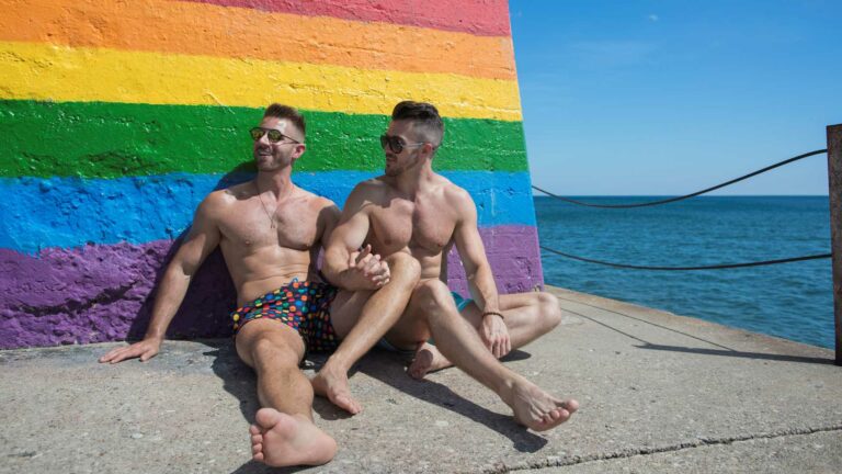 Two men in bathing costumes sit down by a beach holding hands