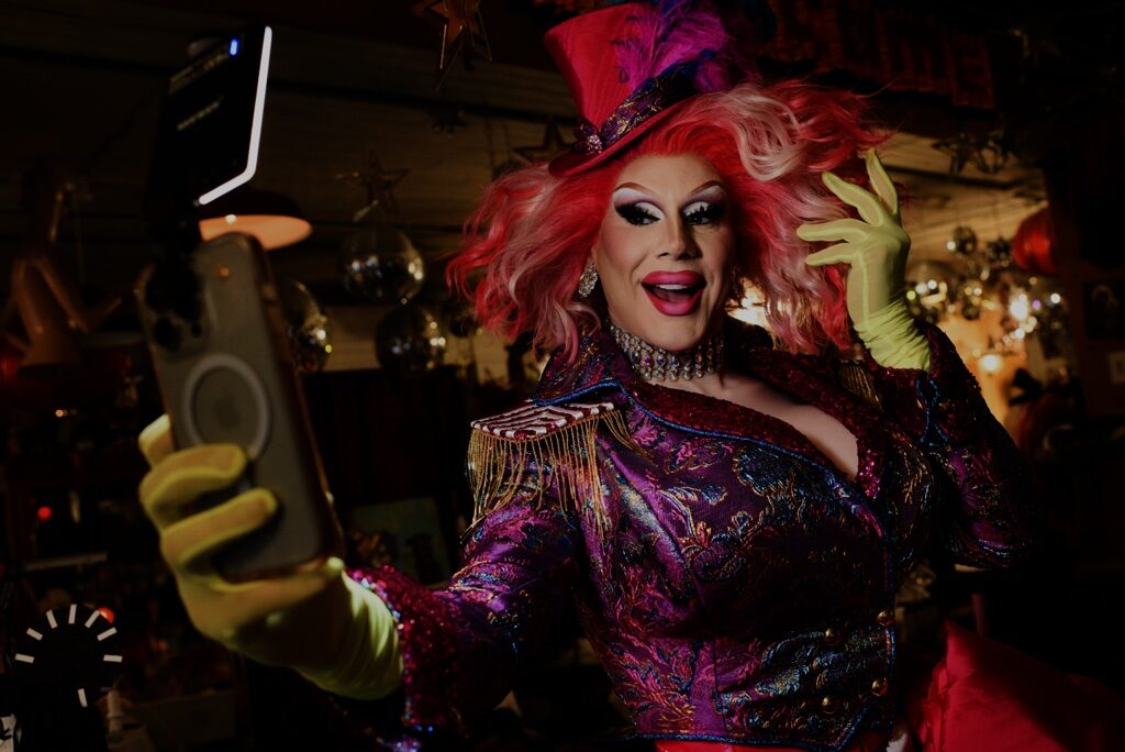 The drag queen Jimbo wearing a pink top hat holding an iPhone taking a selfie of herself