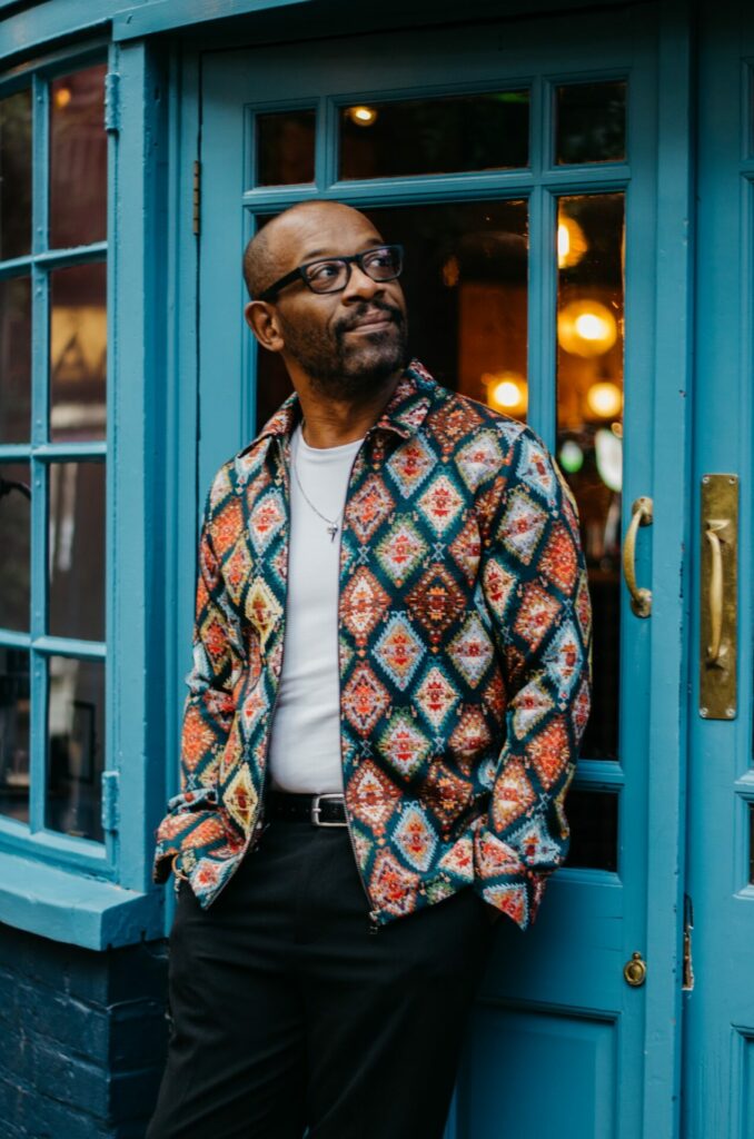 Lennie James wearing a colourful jacket leaning against a blue door