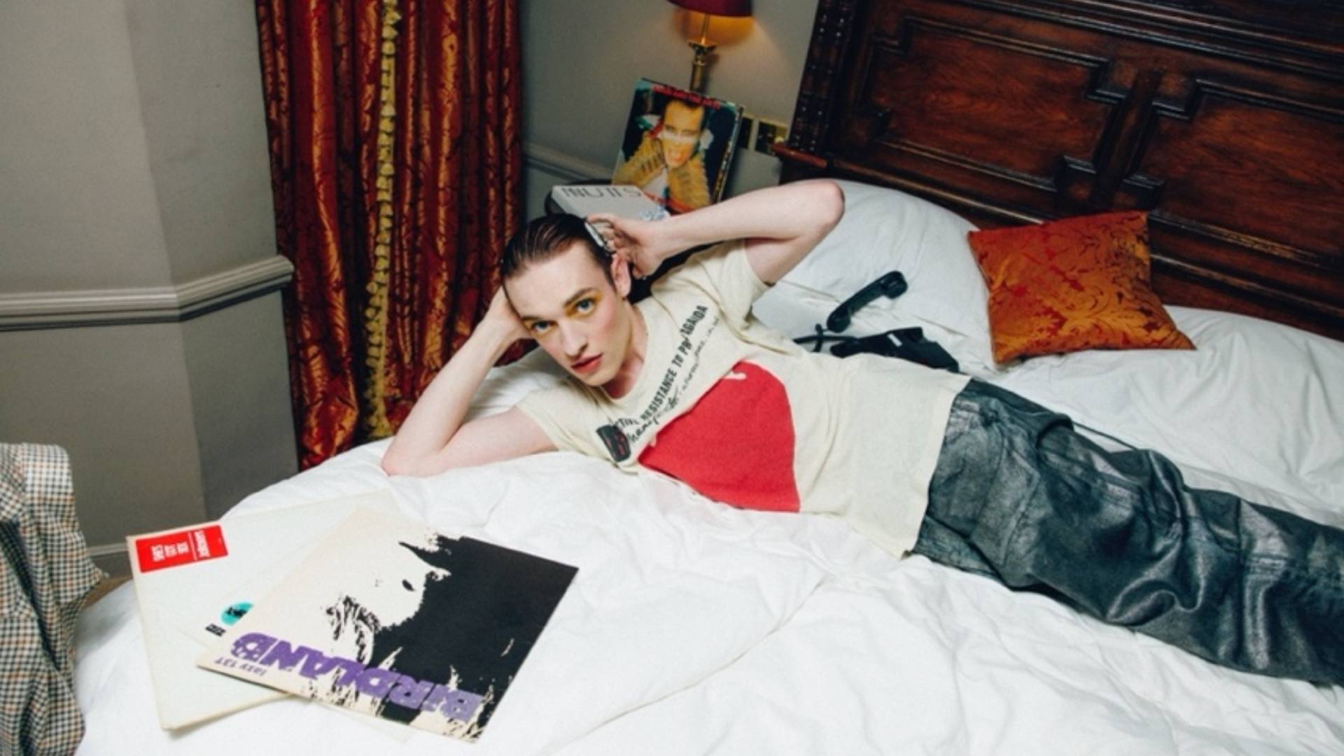 The singer Nemo wearing a white t-shirt with a red logo, lying on a bed