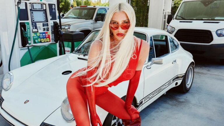 Kesha wearing a red catsuit standing in front of a white car