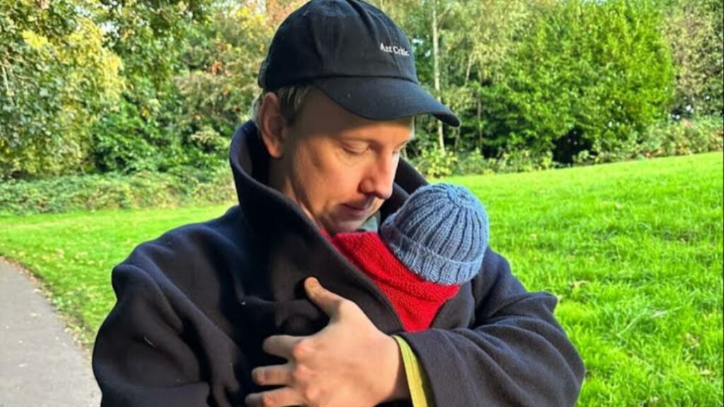 Joe Lycett in a cap cradling a baby