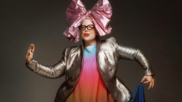 A half-portrait of The Black Madonna wearing a rainbow coloured dress, a silver blazer and a large pink bow in her hair