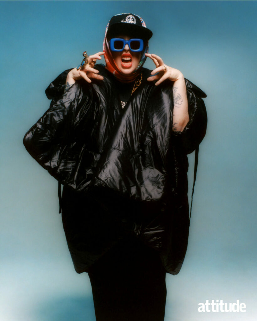 A full-length shot of The Blessed Madonna wearing blue glasses and a black puffer jacket making a zombie pose