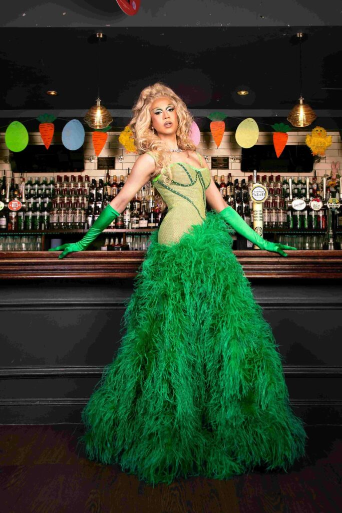 The drag queen Tia Kofi poses in front of a bar while wearing a green dress with a feathered skirt