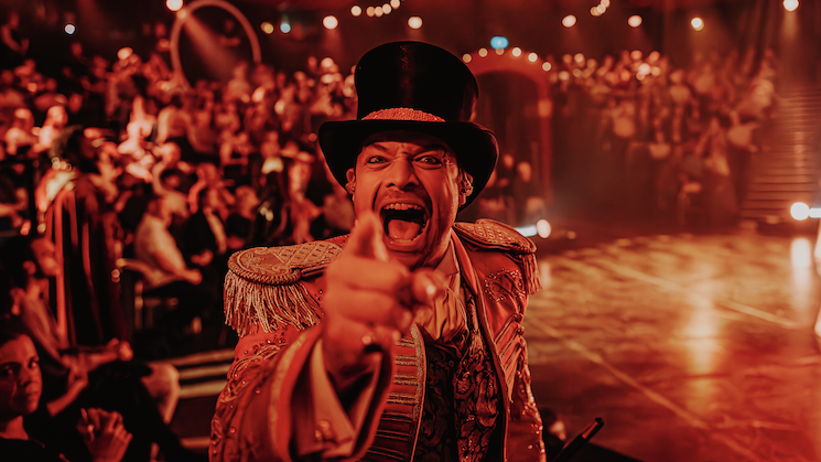 a circus master points at the camera with the audience behind him