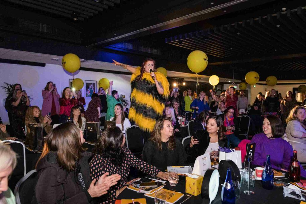 A woman stands dressed as a bee addresses an audience 