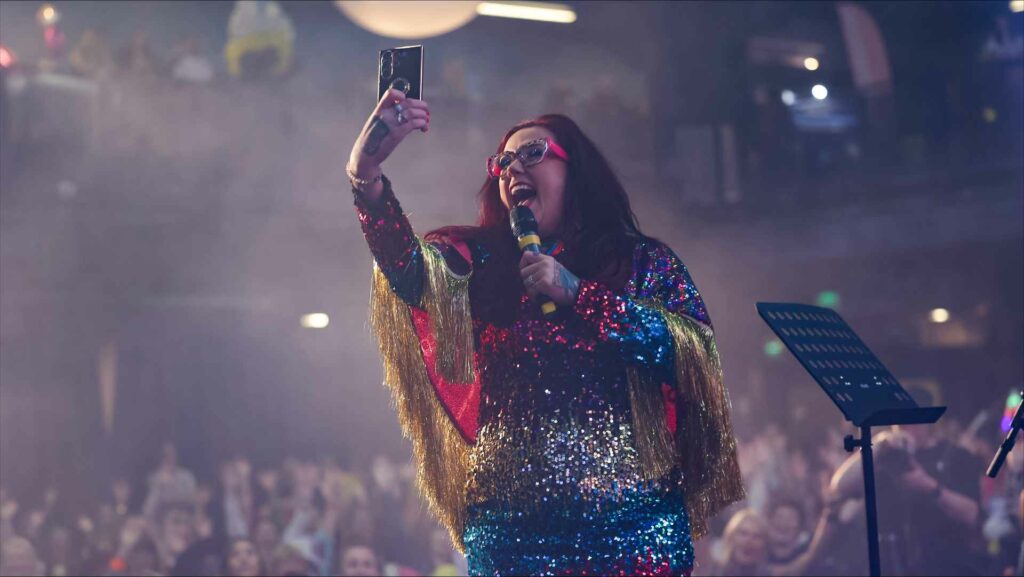 A woman with a mic in her hand on a stage takes a selfie