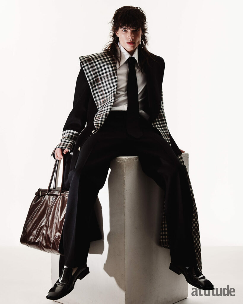 Model sits posing in suit with black and gingham print overcoat 