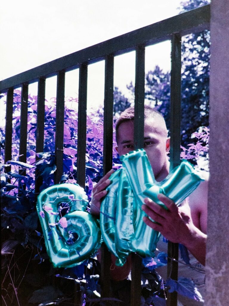 a man standing in a garden by some railings holding some balloons reading 'boy'