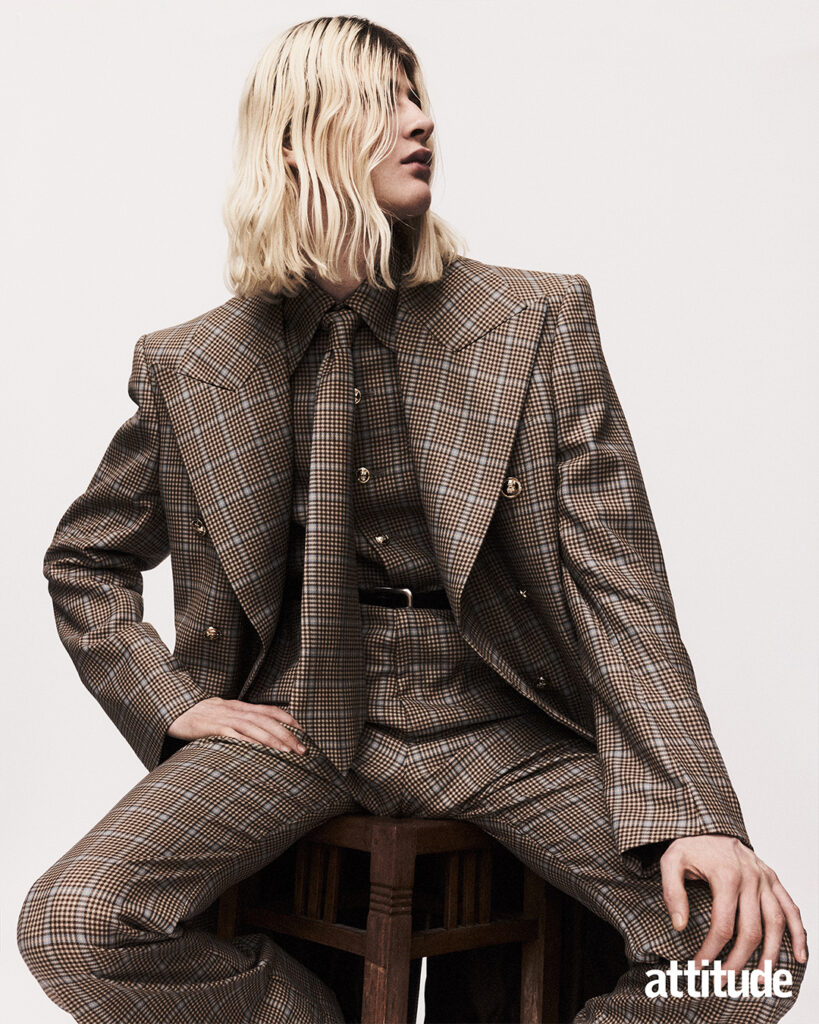 Model sits down in brown check shirt, tie, trousers and overcoat in same fabric