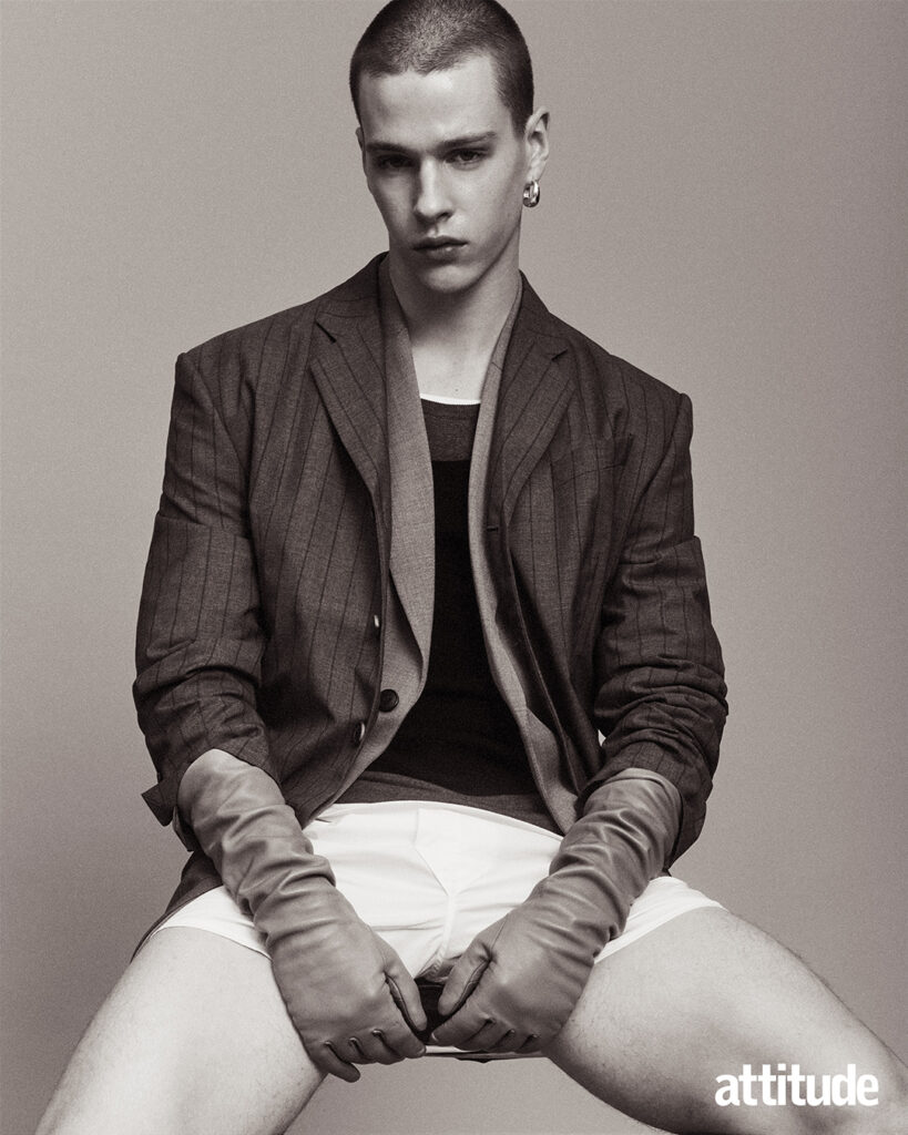 Model poses sitting down in two dark grey blazers, white boxers and leather gloves 