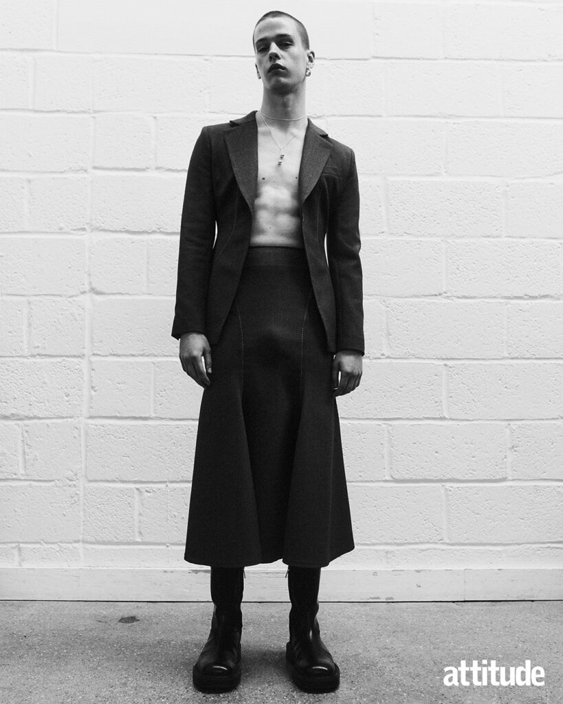 Model stands in coordinating blazer and long skirt with chunky boots and layered necklaces