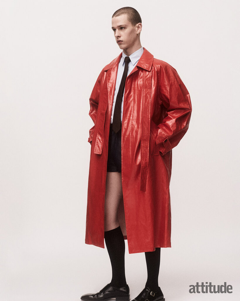 Model poses in red leather trench, white shirt, black tie with long black socks and loafers