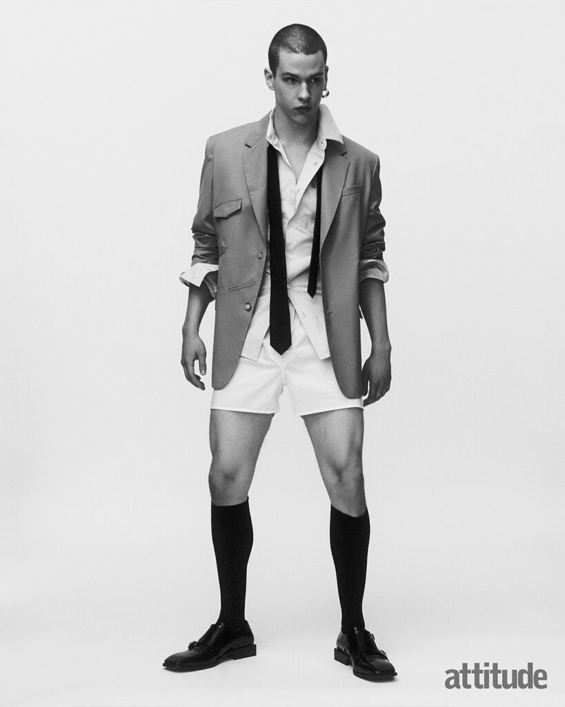 Model poses in open white shirt and boxers, with open tie and blazer with long black socks and loafers