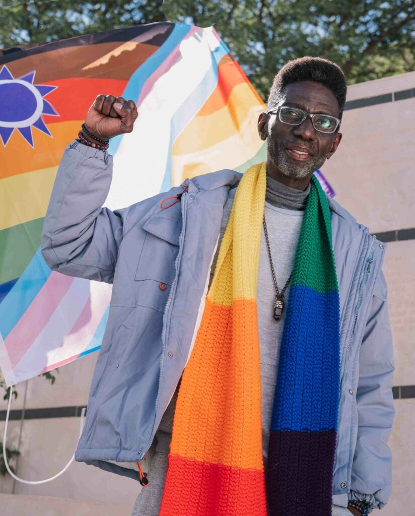 A man wearing a Pride coloured scarve makes a victory fist while looking into the camera