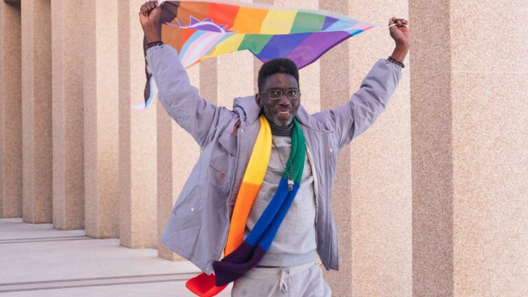 A man in a grey tracksuit holding up a Pride flag over his head with a smile on his face