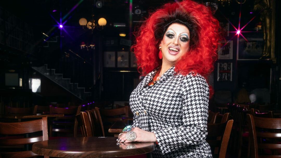 Holly Stars wearing a red wig and black and white dress sitting on a stool in a dark nightclub