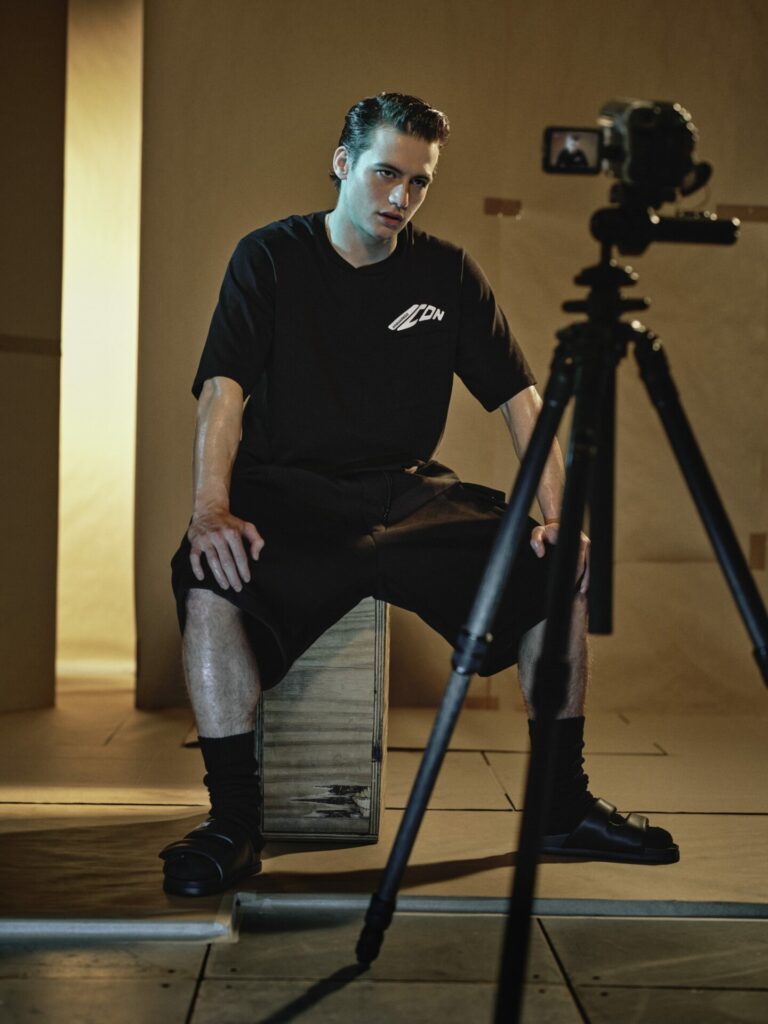 Bách sits in Dsquared's black oversized t-shirt and cargo shorts while posing in front of camcorder on tripod