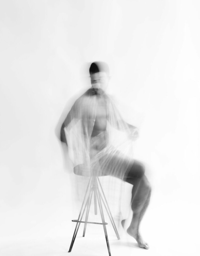 a topless man sitting on a stool draped in white material, out of focus