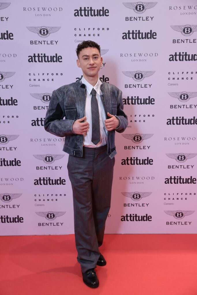 olly Alexander on the red carpet in white shirt, black tie, and grey jacket and trousers