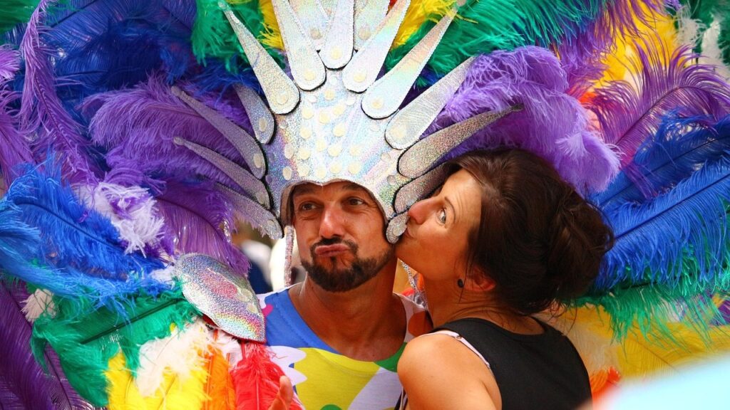 A man in a rainbow feathered headdress is kissed on the cheek by a woman