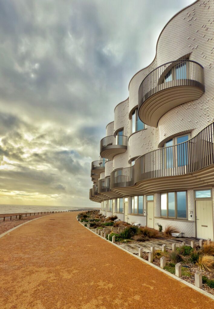 View of an apartment building with white bricks in the shape of waves overlooking the ocean, with a brown path in front of it 