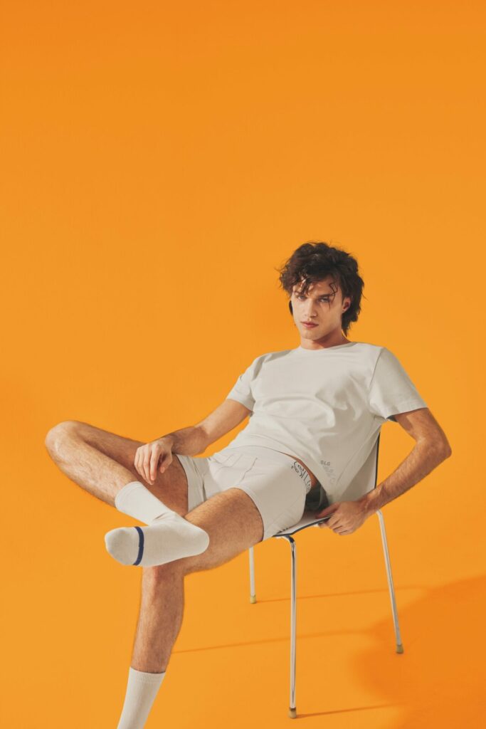 a man in light coloured t-shirt, shorts and socks sat on a chair, orange background