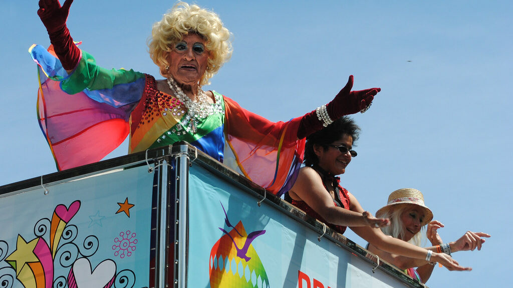 A drag queen in flamboyant clothing stands on a Pride float