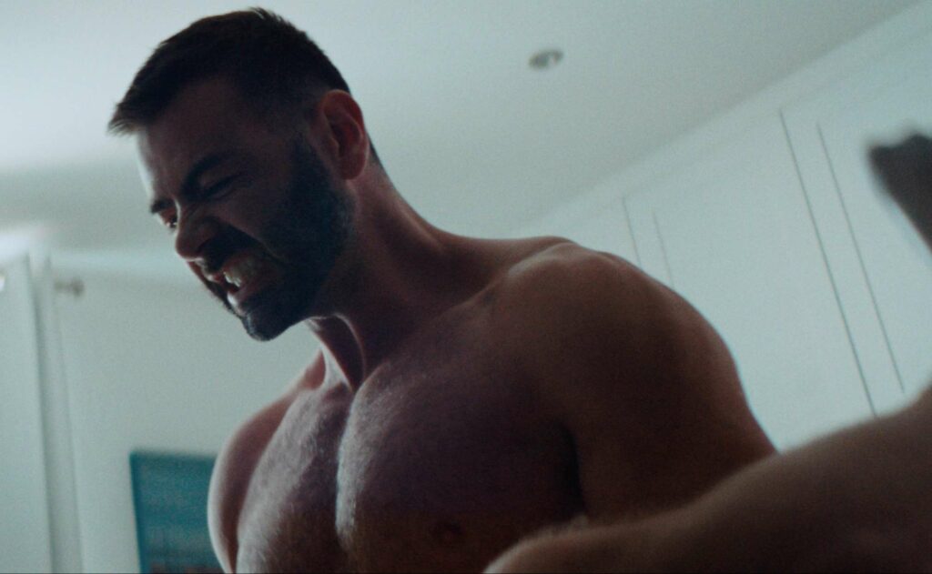 A shirtless, muscular man with a beard clenches his teeth in a brightly lit room