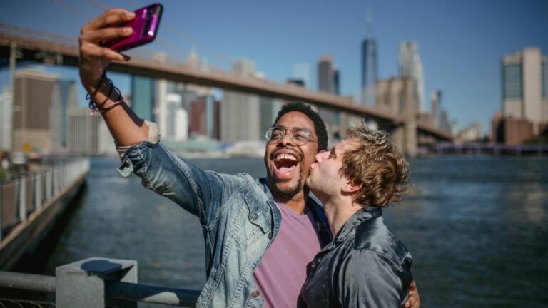 A same-sex couple taking a selfie near the water with the Brooklyn Bridge and New York City skyline in the background.