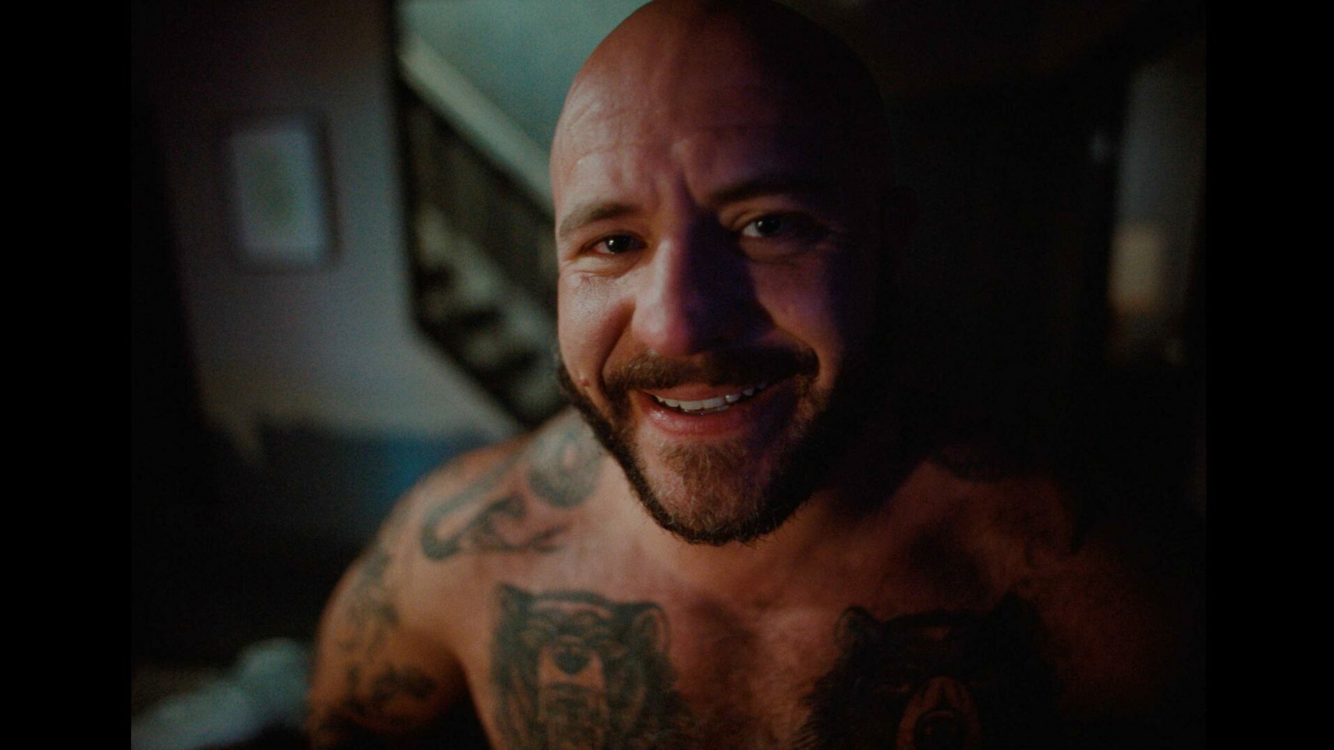 A bald, bearded man with tattoos smiles warmly at the camera in a dimly lit room