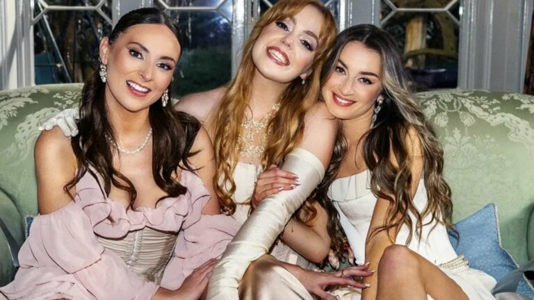 Three women in pastel dresses sit closely together on a vintage-style sofa, smiling at the camera in a room with large windows behind them