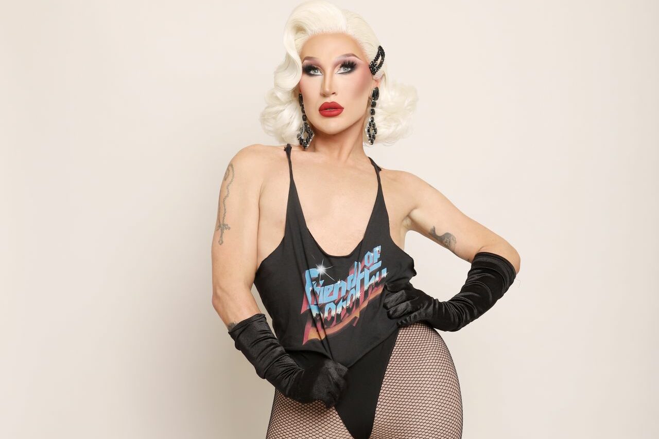 The Vivienne with platinum blonde hair poses in a black tank top, fishnet tights, and gloves