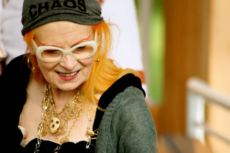 Vivienne Westwood wearing large white glasses, a grey beanie with "CHAOS" written on it, and multiple gold necklaces