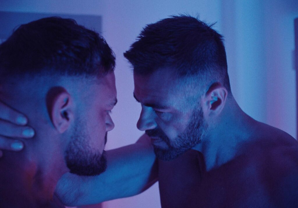 Two shirtless men with beards and short hair face each other closely in a dimly lit room with blue and purple lighting. One man holds the other's neck, their intense eye contact suggesting a moment of intimacy