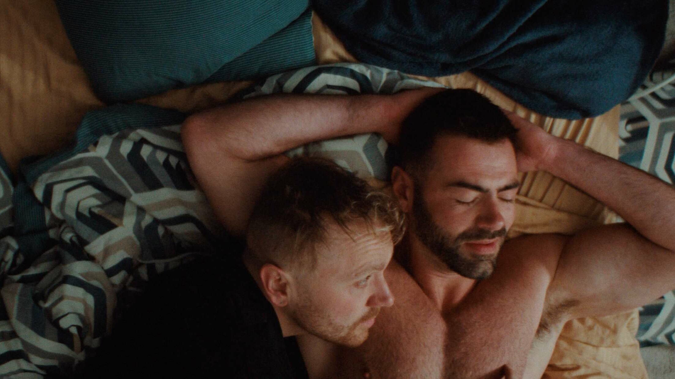 Two men lie together on a bed, one shirtless with his arms behind his head, the other resting close to him