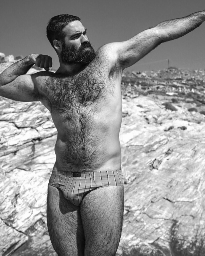 Black and white portrait of Yorgos Tsiantoulas flexing on a beach in speedos